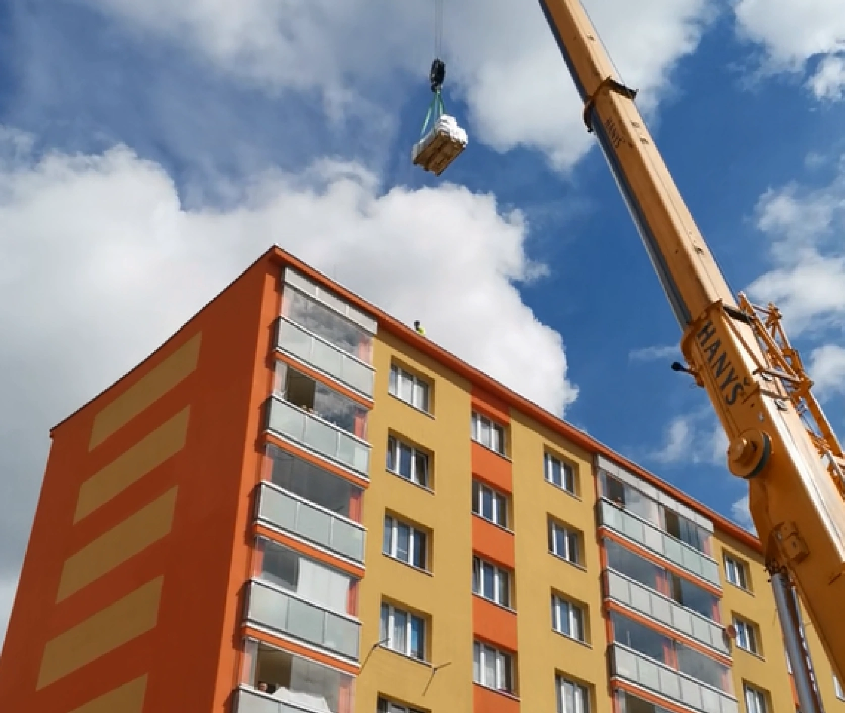 Věžový jeřáb zvedající paletu stavebního materiálu na střechu panelového domu s oranžovo-žlutou fasádou