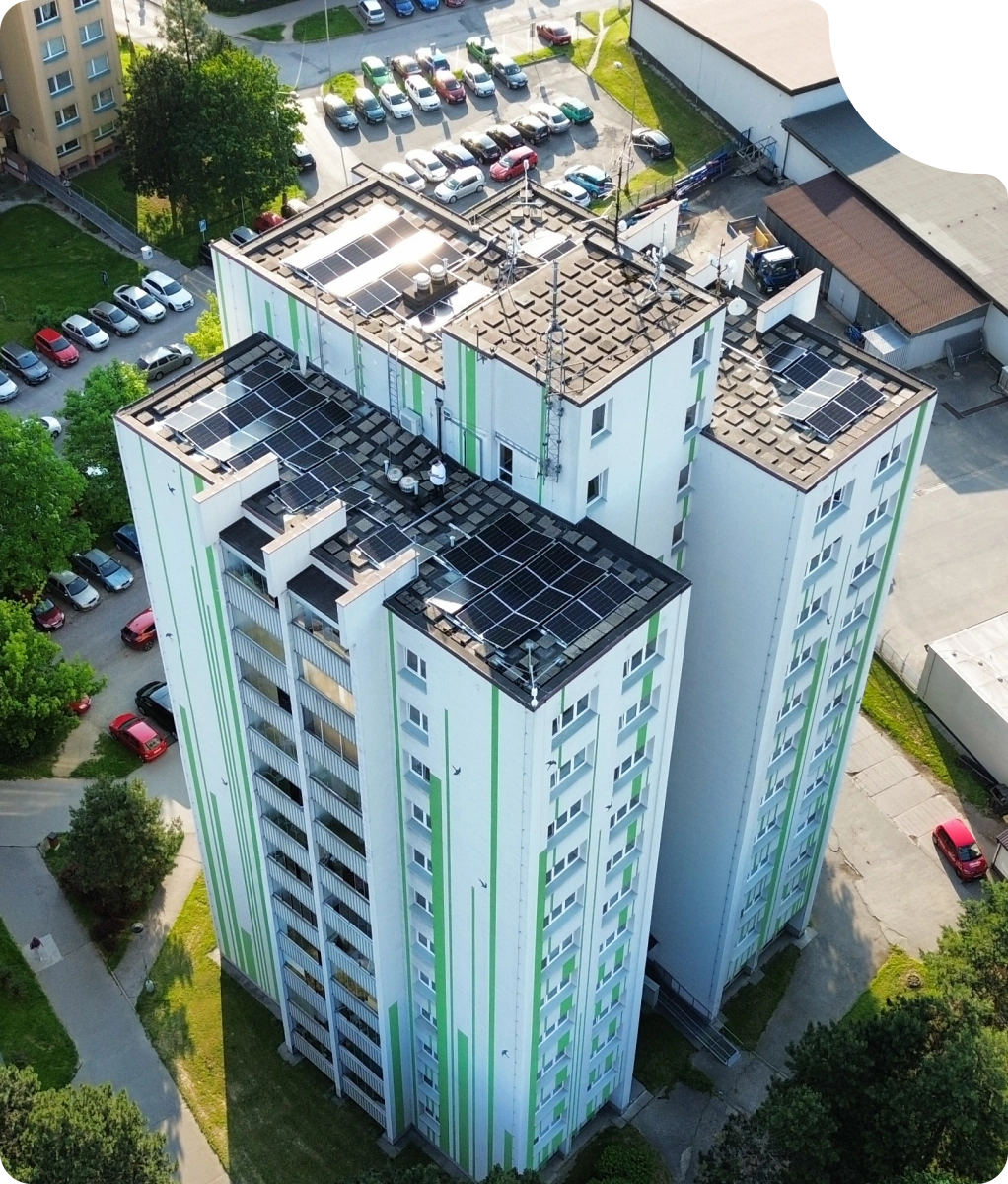 Vysoký bytový dům se solárními panely na střeše, obklopený parkovištěm a stromy